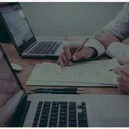 Dwie osoby analizują dokument z obliczeniami przy biurku z dwoma laptopami i długopisami, skupiając się na szczegółach finansowych.