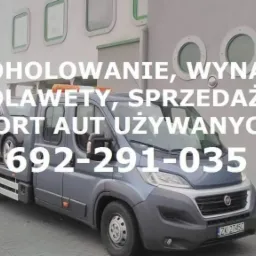 Szara laweta z załadowanym samochodem osobowym na tle budynku z oknami i reklamą kawiarni. Na lawecie widoczne pomarańczowe światła ostrzegawcze.