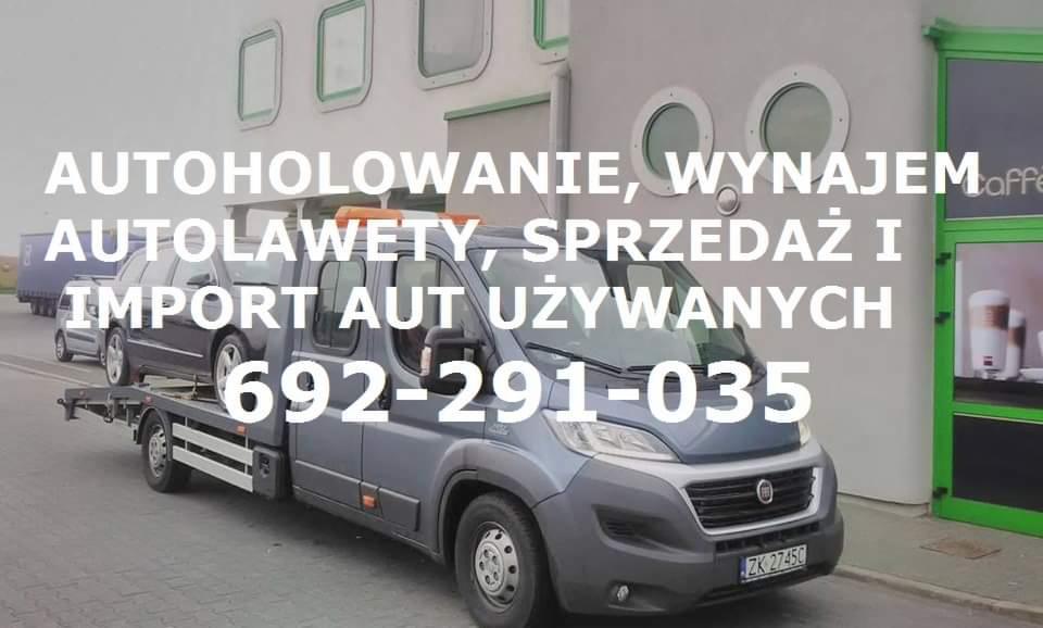 Szara laweta z załadowanym samochodem osobowym na tle budynku z oknami i reklamą kawiarni. Na lawecie widoczne pomarańczowe światła ostrzegawcze.