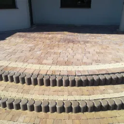 Wykończony taras z kostki brukowej w odcieniach brązu i beżu, z półokrągłymi schodami i dekoracyjnym obramowaniem z drobnego żwiru, przylegający do jasnego budynku.