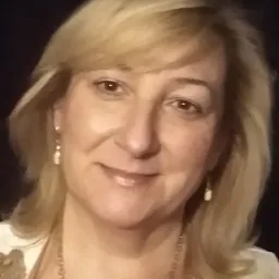 Portret uśmiechniętej kobiety z blond włosami, noszącej złoty łańcuszek i kolczyki w kształcie łezki, na ciemnym tle.