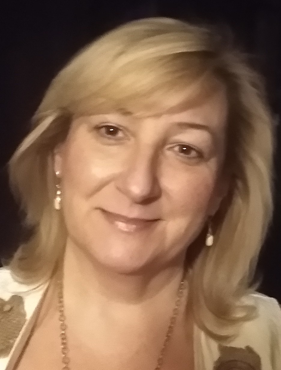 Portret uśmiechniętej kobiety z blond włosami, noszącej złoty łańcuszek i kolczyki w kształcie łezki, na ciemnym tle.