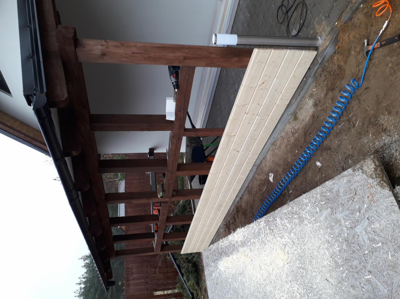 Drewniana konstrukcja z widoczną więźbą dachową, częściowo obita boazerią, z narzędziami w tle i niebieskim wężem pneumatycznym na ziemi.