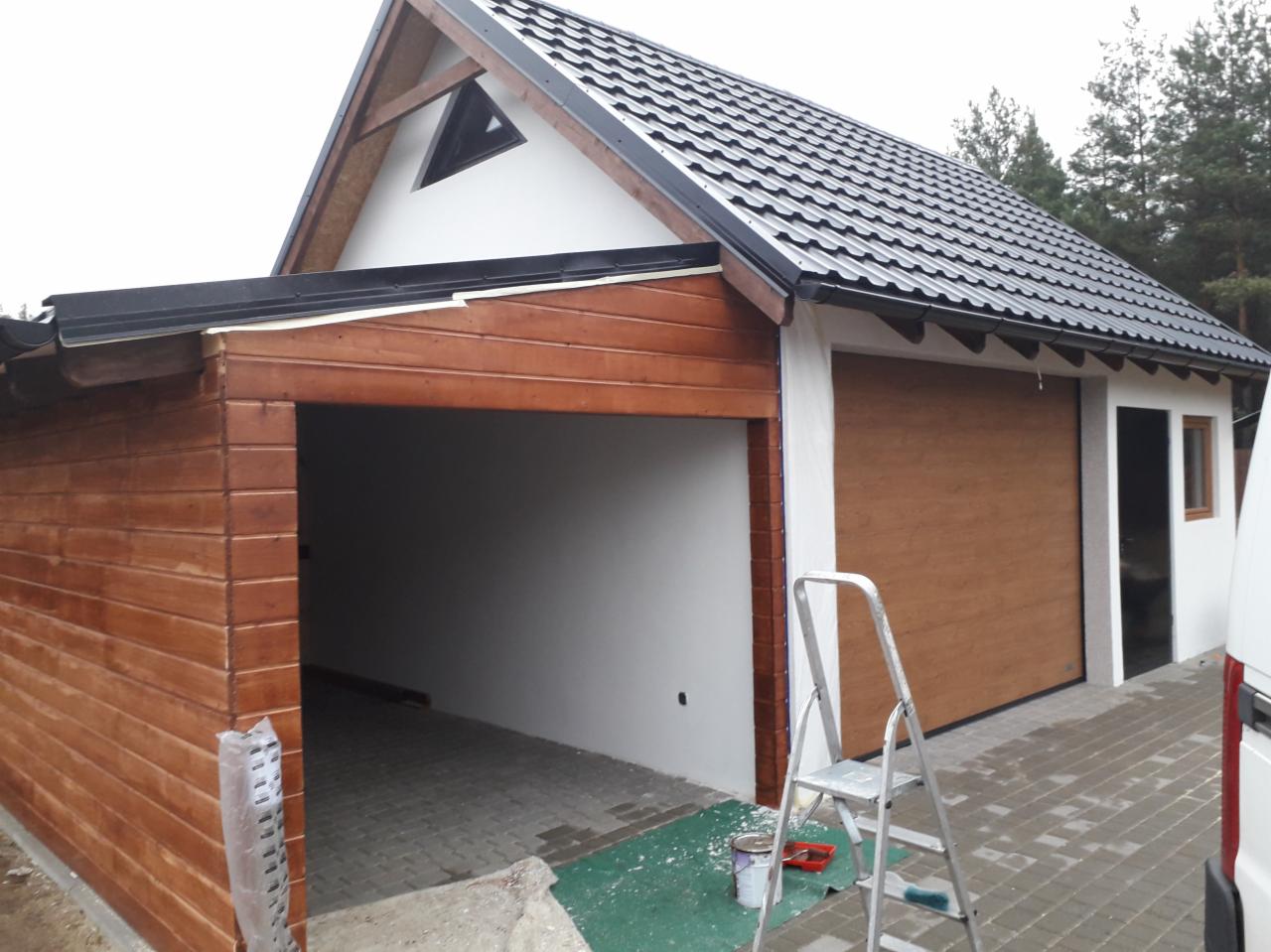 Garaż wolnostojący z drewnianą elewacją i brązową bramą segmentową, częściowo pomalowany na biało, z metalową drabinką opartą o ścianę, widoczne ślady farby i puszka z farbą, dach pokryty...