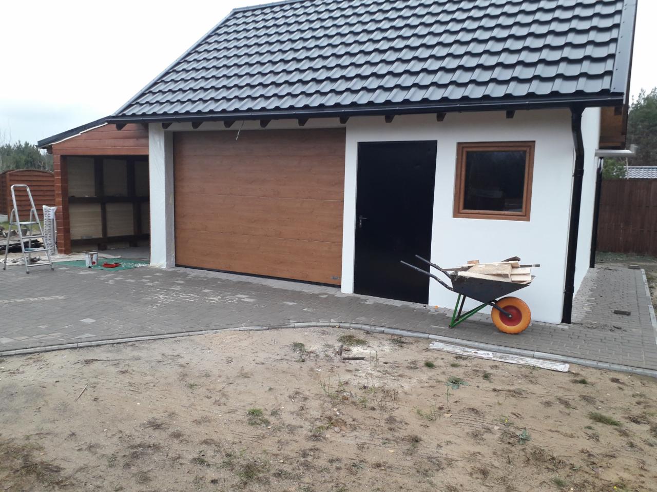 Nowoczesny garaż z brązową bramą segmentową, czarnymi drzwiami wejściowymi i oknem w drewnianej ramie, obok taczka z drewnem, kostka brukowa przed budynkiem.