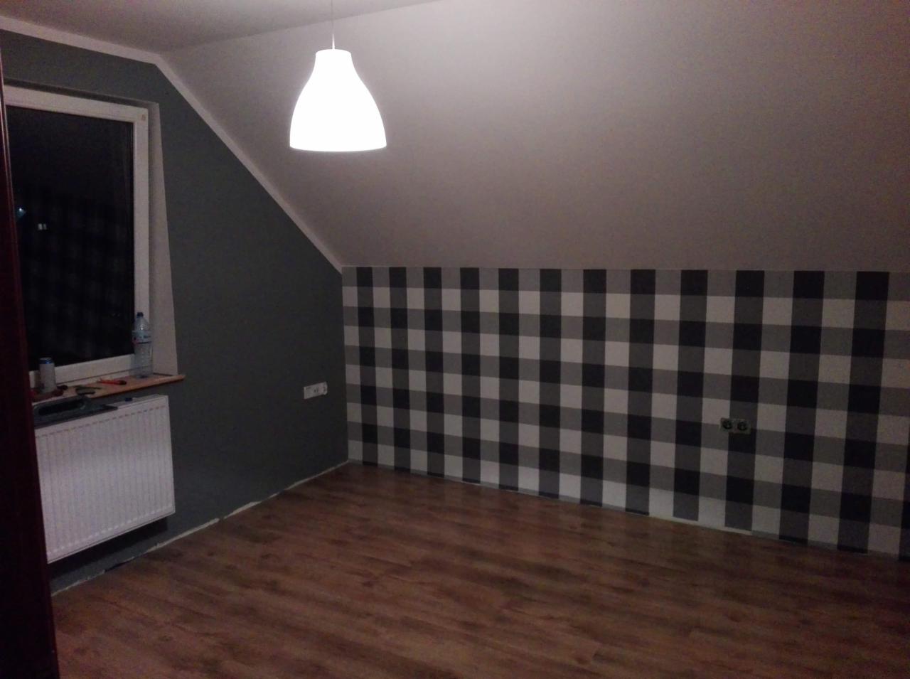 Poddasze po odświeżeniu: ściana w czarno-białą kratę, szara ściana z oknem, drewniana podłoga, biały grzejnik i lampa sufitowa.