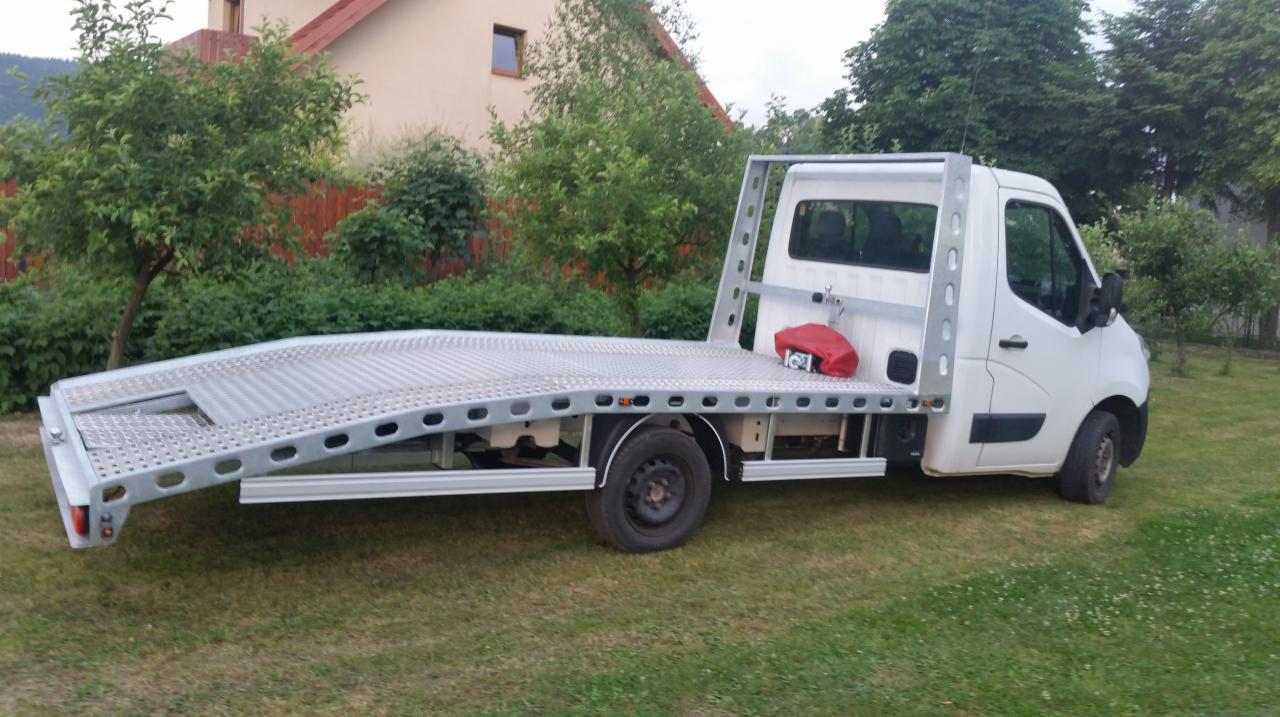 Biała laweta z aluminiową platformą załadunkową, zaparkowana na trawniku przed domem z czerwonym dachem.