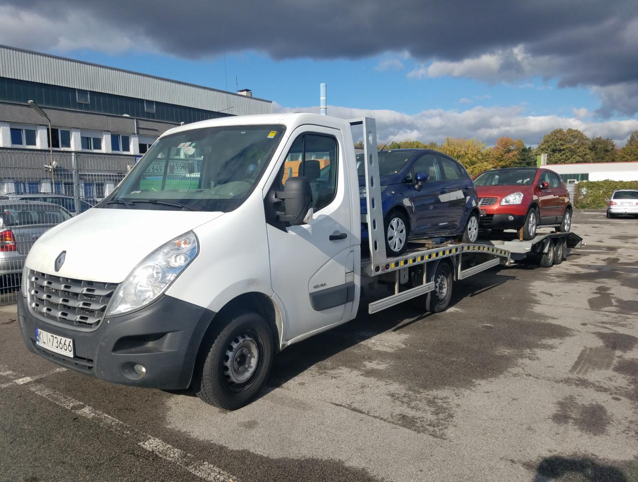 Biała laweta Renault Master z dwoma samochodami (niebieskim i czerwonym) na platformie, zaparkowana na parkingu z widocznym budynkiem w tle, pochmurne niebo.