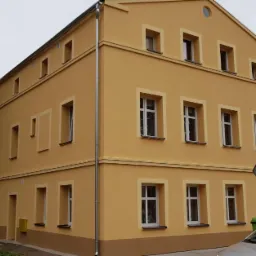 Odnowiona elewacja budynku dwupiętrowego w odcieniach żółci i beżu, z nowymi oknami i rynnami, widok z ulicy w pochmurny dzień.