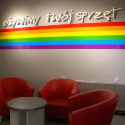 Poczekalnia firmy IT w Szczecinie z czerwonymi fotelami i okrągłym stolikiem, na ścianie napis 'ożywimy twój sprzęt' nad tęczowym paskiem.