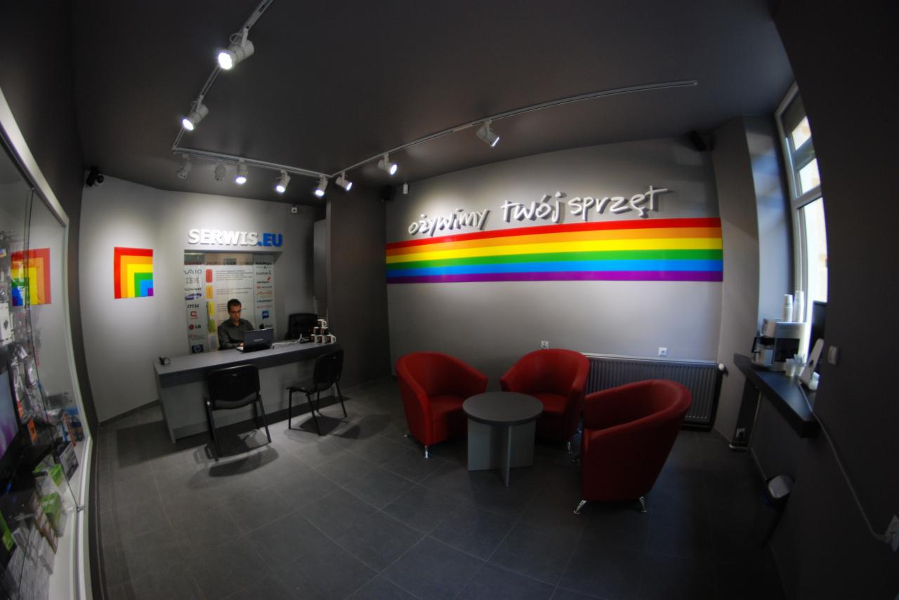 Wnętrze punktu serwisowego komputerów z recepcją, stanowiskiem pracy z laptopem, miejscem oczekiwania z czerwonymi fotelami i tęczowym logotypem 'ożywimy twój sprzęt' na ścianie.
