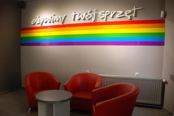 Poczekalnia firmy IT w Szczecinie z czerwonymi fotelami i okrągłym stolikiem, na ścianie napis 'ożywimy twój sprzęt' nad tęczowym paskiem.