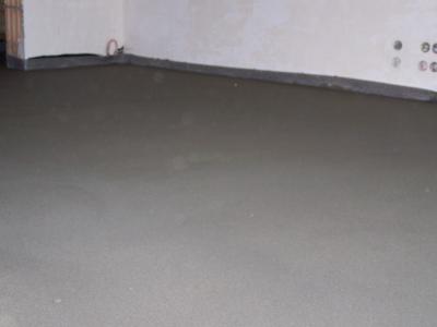 Świeżo wylana, gładka posadzka cementowa w pomieszczeniu z surowymi ścianami i widocznymi gniazdkami elektrycznymi, gotowa do dalszych prac wykończeniowych.