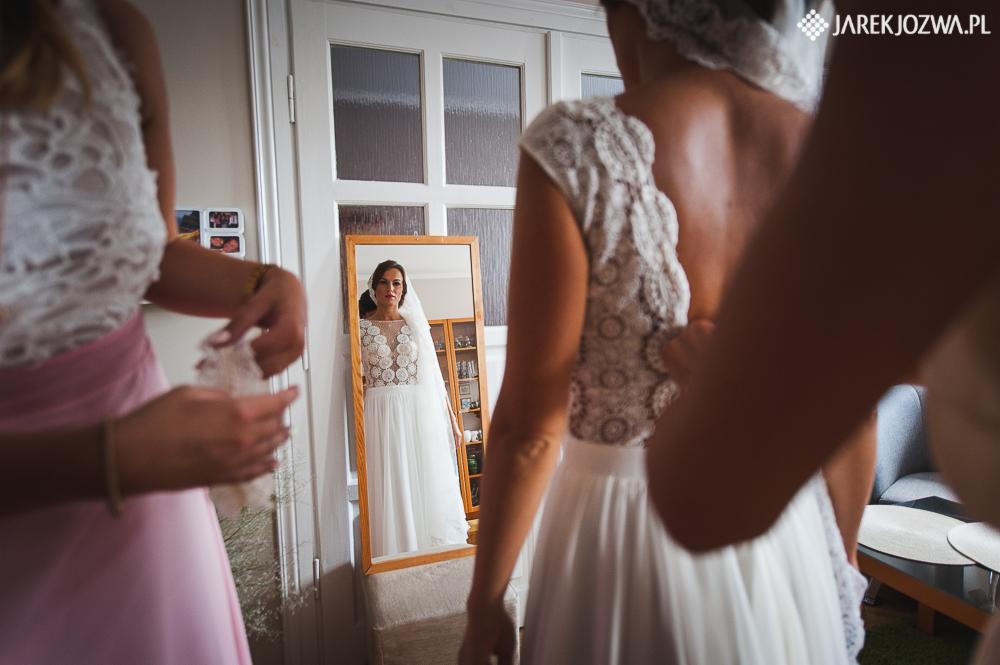 Panna młoda w sukni z koronkową górą, widziana w lustrze podczas przygotowań ślubnych, asystuje jej osoba w różowej sukience.