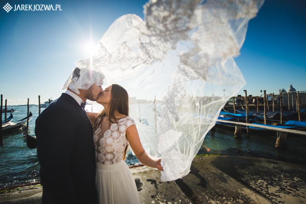 Para młoda całuje się w Wenecji, zasłonięta delikatnym, koronkowym welonem na tle gondoli i błękitnego nieba.