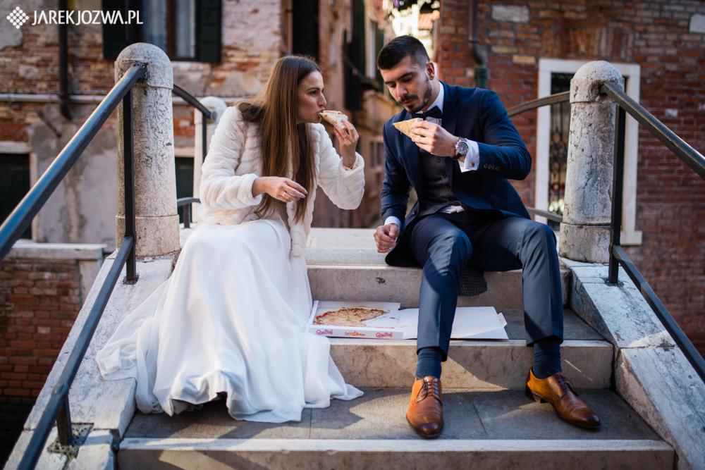 Para młoda w strojach ślubnych siedzi na schodach mostu w Wenecji, jedząc pizzę z kartonowego pudełka. Panna młoda w futrzanym bolerku. Pan młody w granatowym garniturze z muchą.