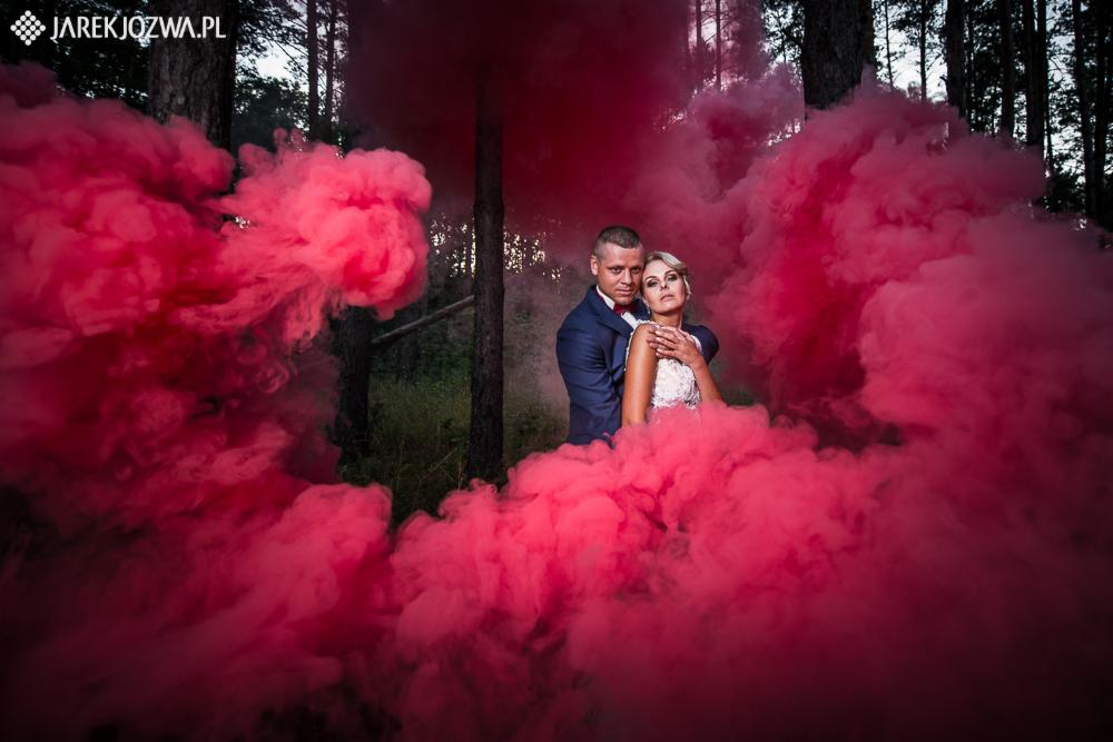 Para młoda w lesie otoczona czerwonym dymem, on w granatowym garniturze, ona w białej sukni ślubnej z koronką, oboje patrzą w obiektyw.