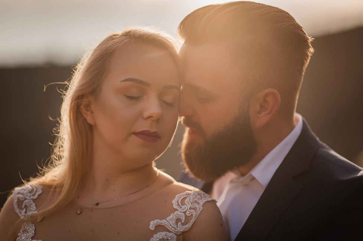 Para młoda w blasku zachodzącego słońca, panna młoda w sukni z koronkowymi aplikacjami na ramionach, pan młody w ciemnym garniturze, oboje z zamkniętymi oczami.