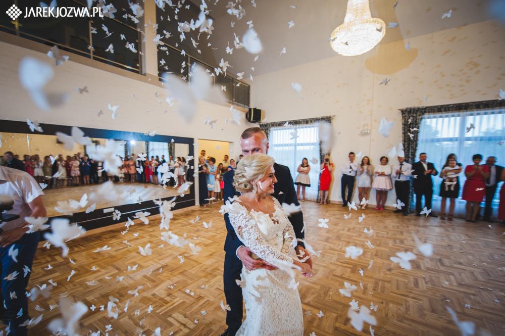 Para młoda w tańcu, obsypana białymi papierowymi motylami w przestronnej sali balowej z drewnianą podłogą i lustrami na ścianach, goście obserwują.