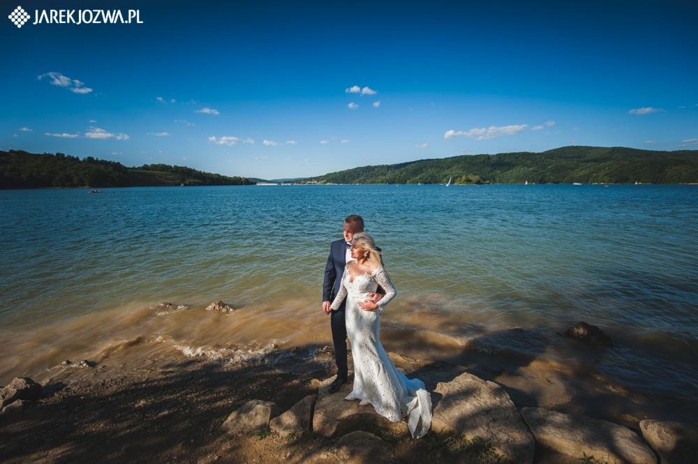 Para młoda w strojach ślubnych stoi na skalistym brzegu jeziora w słoneczny dzień, z widokiem na zalesione wzgórza w tle.
