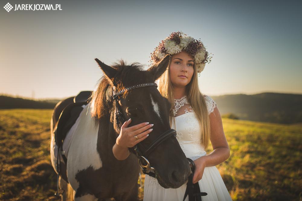 Panna młoda w białej sukni i wianku pozuje z koniem na tle łąki w ciepłym świetle zachodzącego słońca.