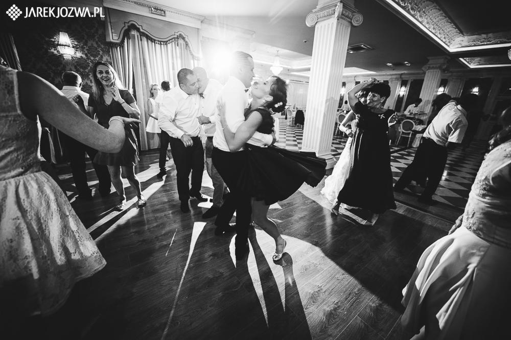 Czarno-białe zdjęcie z wesela: para tańczy na parkiecie, otoczona gośćmi w eleganckim wnętrzu z kolumnami i wzorzystą podłogą. Długie cienie i jasne światło tworzą dynamiczną atmosferę.