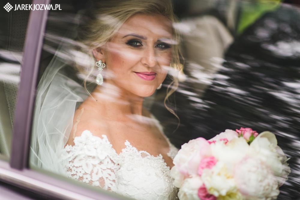 Panna młoda w sukni z koronkowym dekoltem, z bukietem białych i różowych piwonii, widziana przez szybę samochodu z refleksami światła.