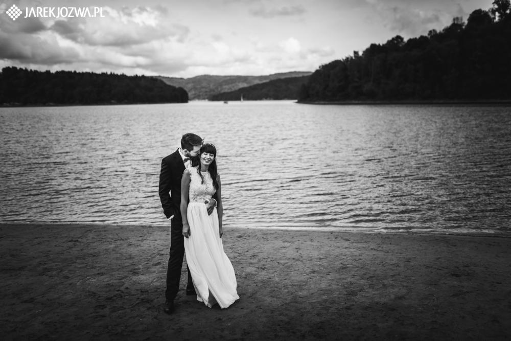 Czarno-białe zdjęcie pary młodej na plaży, z jeziorem i zalesionymi wzgórzami w tle; pan młody w czarnym garniturze całuje pannę młodą w głowę, panna młoda w białej sukni z koronkową górą uśmiecha...