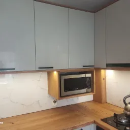 Nowoczesna kuchnia z szarymi szafkami wiszącymi z drewnianymi akcentami, mikrofalówką Samsung w drewnianej wnęce, blatem z jasnego drewna i marmurową ścianą. Na blacie czajnik na płycie grzewczej.