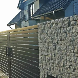 Nowoczesna brama wjazdowa z poziomych profili stalowych, zamontowana pomiędzy kamiennymi słupkami z jasnego granitu, na tle domu z ciemną dachówką.