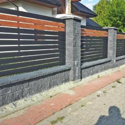 Pro-Moderno - Nowoczesne ogrodzenie panelowe z poziomymi, grafitowymi przęsłami i elementami imitującymi drewno, osadzone na słupkach i murku z szarej kostki łupanej.