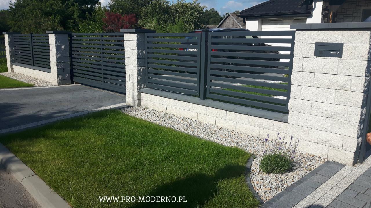 Nowoczesne ogrodzenie panelowe z szarej stali na podmurówce z jasnego kamienia łupanego, z wbudowaną skrzynką na listy, otoczone trawnikiem i ozdobnym kamieniem.