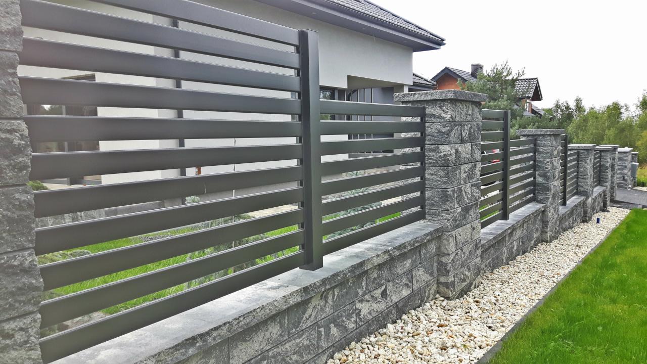 Nowoczesne ogrodzenie panelowe z poziomych, szarych profili, osadzone na murku z kamienia łupanego i słupkach z tego samego materiału, z widoczną zielenią trawnika i białym żwirem.