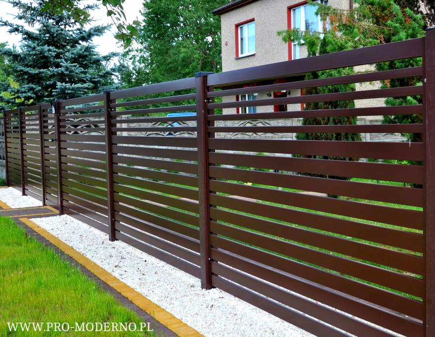Nowoczesne, brązowe ogrodzenie panelowe z poziomych listew, widoczne z boku, z białym żwirem u podstawy i budynkiem w tle.