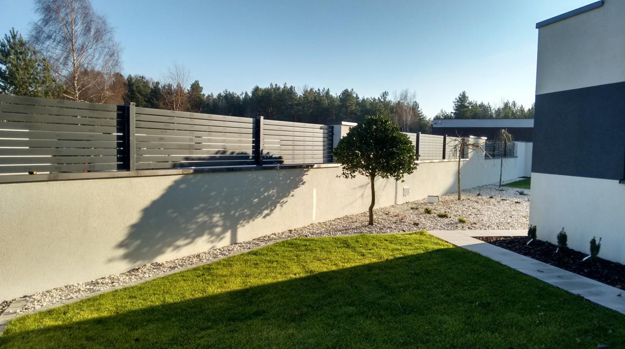 Nowoczesne ogrodzenie panelowe w kolorze antracytowym, zamontowane na niskim murku, z widocznym trawnikiem i młodymi drzewkami w ogrodzie.