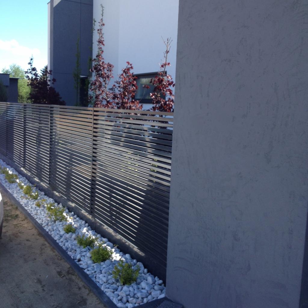 Nowoczesne ogrodzenie z poziomych, metalowych listew w kolorze antracytowym, ozdobione białymi kamieniami i niskimi krzewami, na tle minimalistycznego domu.