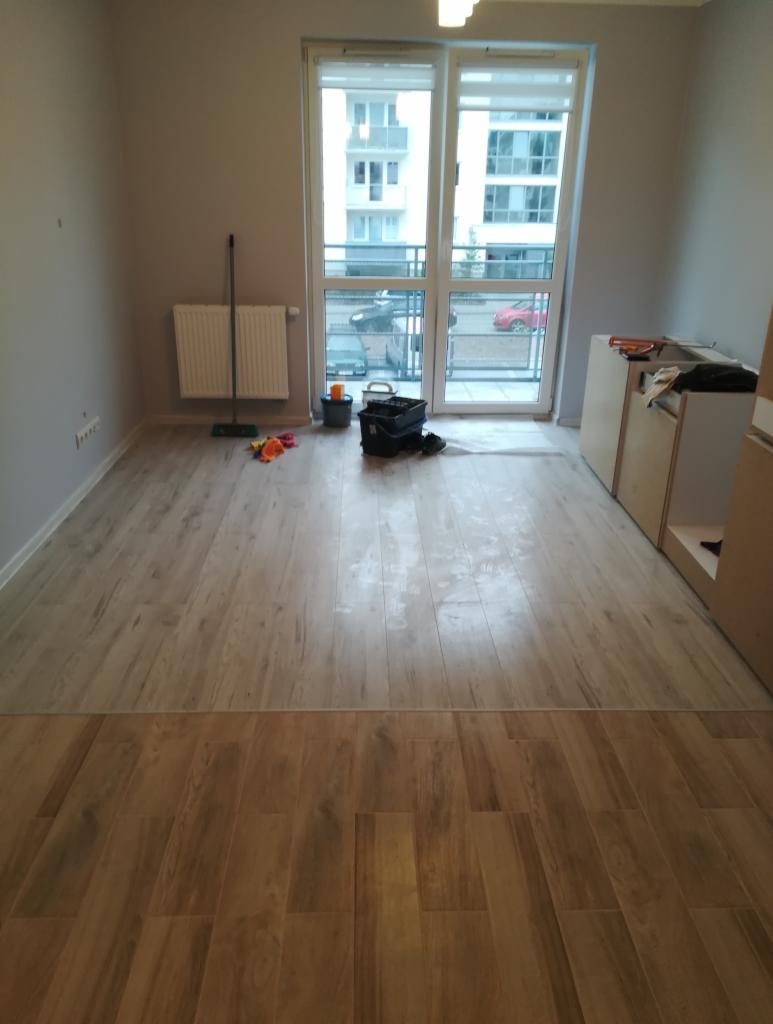 Nowo położona podłoga z paneli drewnopodobnych w jasnym odcieniu przechodząca w ciemniejszy odcień, widoczne narzędzia i akcesoria glazurnicze przy oknie balkonowym, fragment zabudowy kuchennej po...