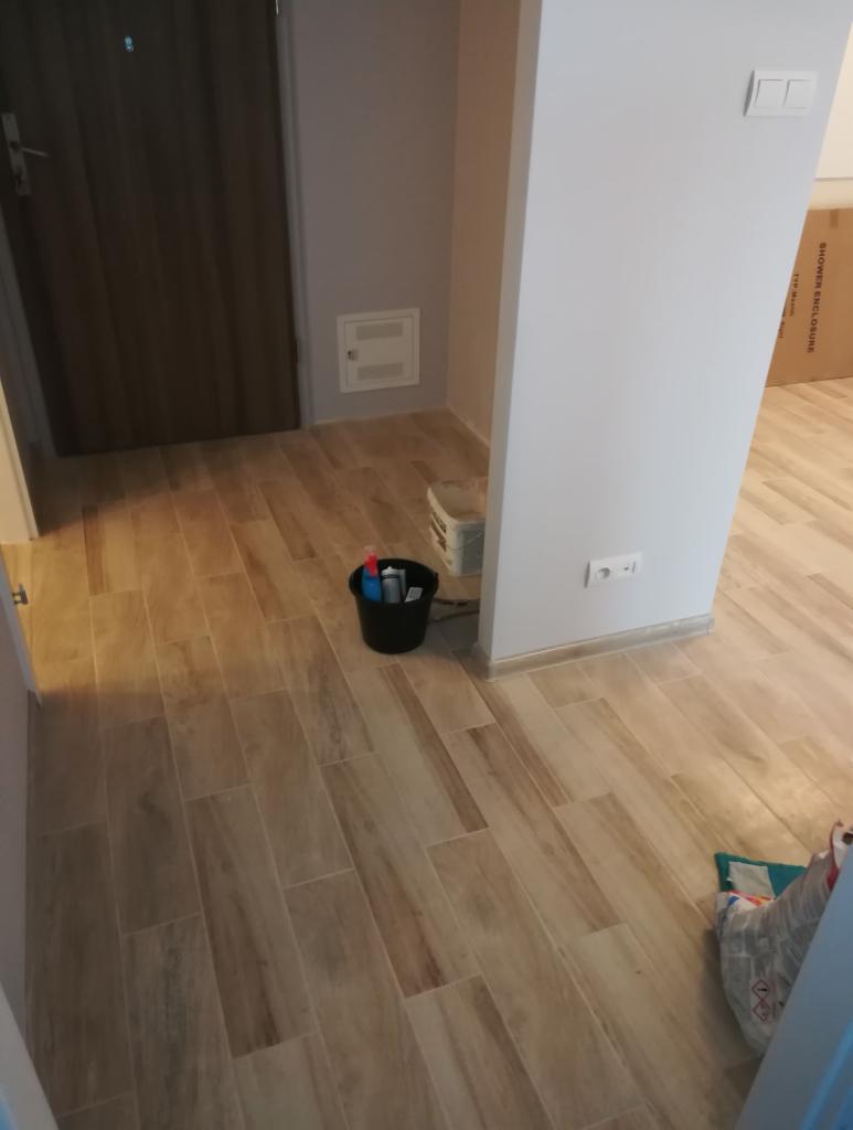 Ułożona podłoga z płytek imitujących drewno w przedpokoju, widoczne wiadro z akcesoriami do czyszczenia, fragment kartonu i drzwi wejściowe.