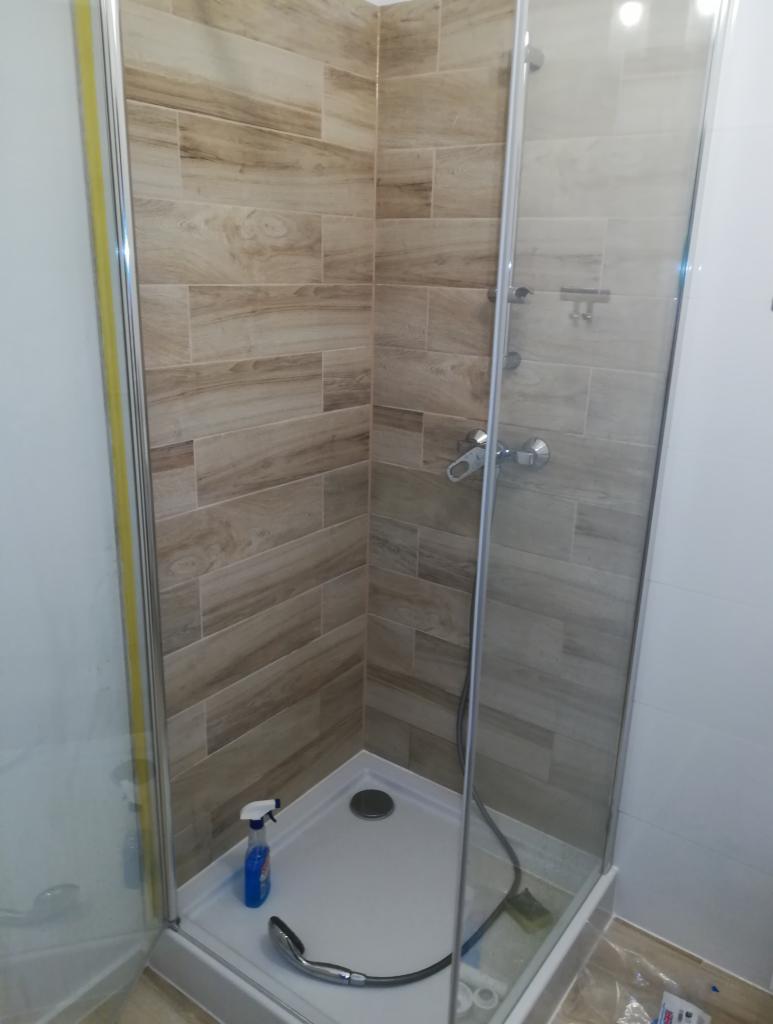 Kabina prysznicowa z beżowymi płytkami imitującymi drewno, widoczny brodzik, bateria i słuchawka prysznicowa, z boku butelka środka czyszczącego.