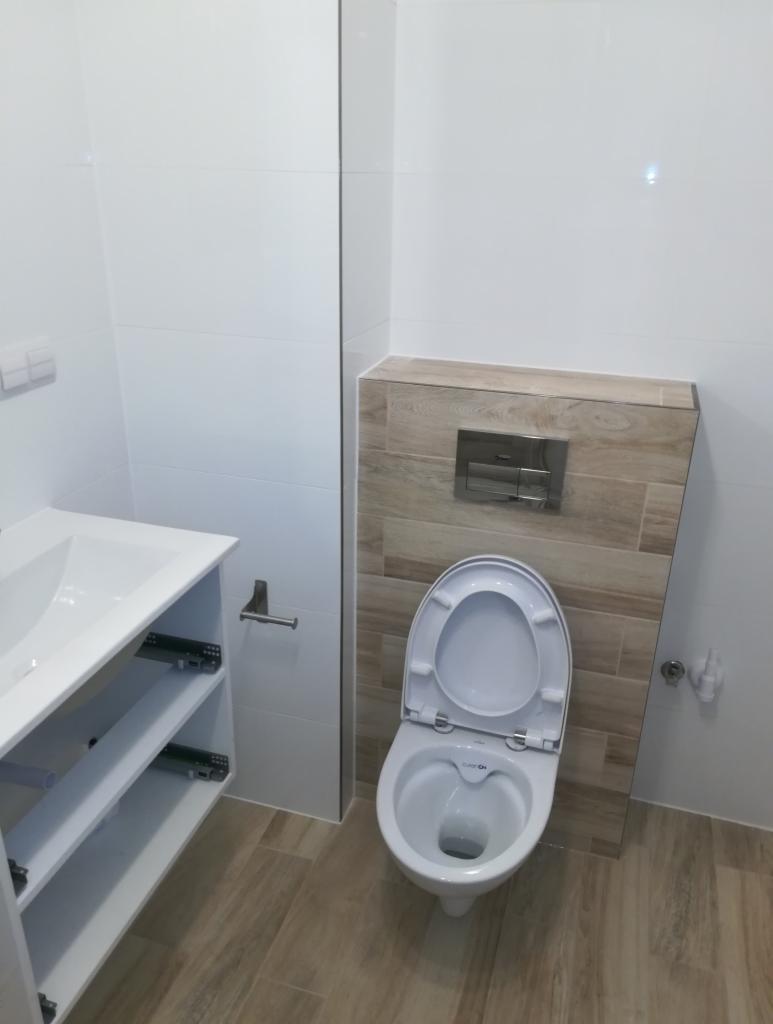 Nowoczesna łazienka z białymi płytkami ściennymi i beżowymi płytkami podłogowymi, wisząca toaleta z zabudowaną spłuczką, biała szafka pod umywalką z otwartymi półkami.