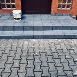 Wykończone schody wejściowe z szarych płytek granitowych, prowadzące do brązowych drzwi, z widoczną puszką farby na górnym stopniu i kostką brukową przed schodami.