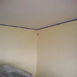 Żółta ściana w trakcie malowania, zabezpieczona niebieską taśmą malarską przy suficie, widoczny fragment drabiny zabezpieczonej folią malarską.