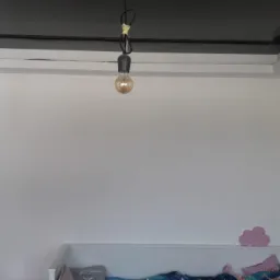 Pokój dziecięcy z czarnym, błyszczącym sufitem podwieszanym, białe ściany, jedna z tapetą w delikatny wzór, wisząca żarówka na kablu, widoczny fragment łóżeczka dziecięcego.