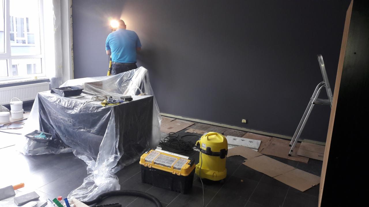 Pracownik na drabinie maluje ścianę na ciemny kolor, w pomieszczeniu zabezpieczone meble folią, widoczny odkurzacz przemysłowy i skrzynka z narzędziami.