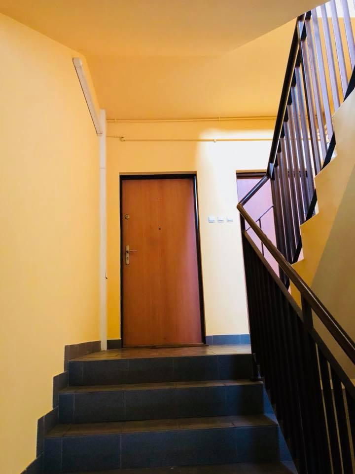 Klatka schodowa w budynku wielorodzinnym po odświeżeniu, widoczne odmalowane ściany w odcieniach żółci, drewniane drzwi wejściowe i ciemne, kamienne schody z metalową balustradą.