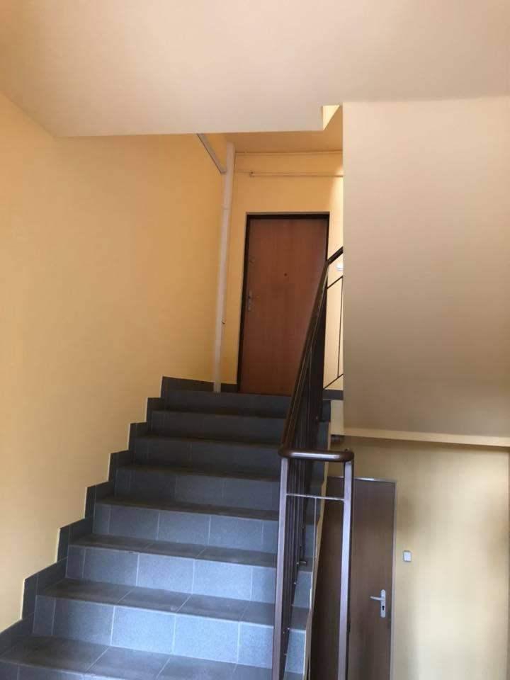 Klatka schodowa po odświeżeniu, ściany pomalowane na żółto, szare płytki na schodach, brązowa poręcz i drzwi.