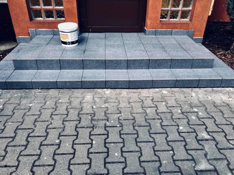 Wykończone schody wejściowe z szarych płytek granitowych, prowadzące do brązowych drzwi, z widoczną puszką farby na górnym stopniu i kostką brukową przed schodami.