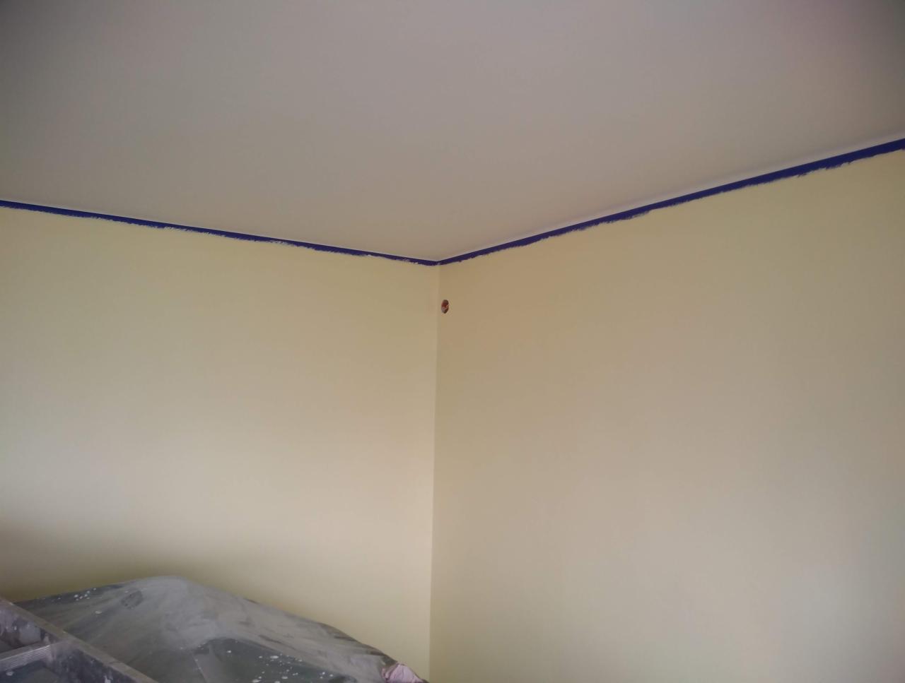 Żółta ściana w trakcie malowania, zabezpieczona niebieską taśmą malarską przy suficie, widoczny fragment drabiny zabezpieczonej folią malarską.