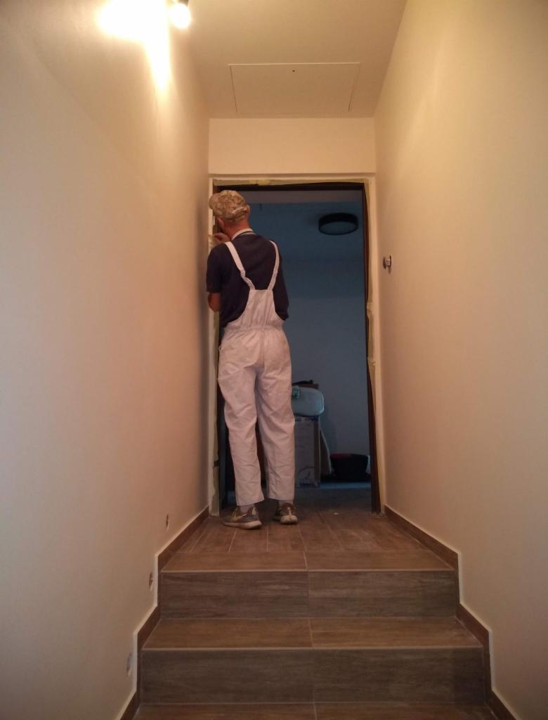 Mężczyzna w białym kombinezonie malarskim zabezpiecza taśmą malarską ościeżnicę drzwi w wąskim korytarzu wykończonym płytkami imitującymi drewno.