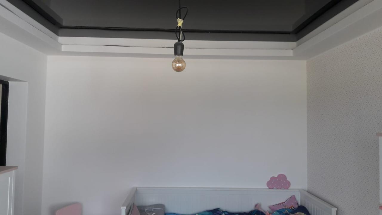 Pokój dziecięcy z czarnym, błyszczącym sufitem podwieszanym, białe ściany, jedna z tapetą w delikatny wzór, wisząca żarówka na kablu, widoczny fragment łóżeczka dziecięcego.